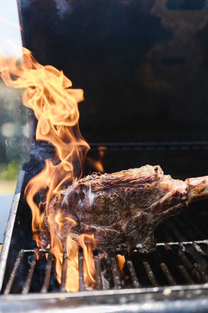 A sizzling tomahawk steak on a barbecue grill with vibrant flames during a sunny outdoor cookout.